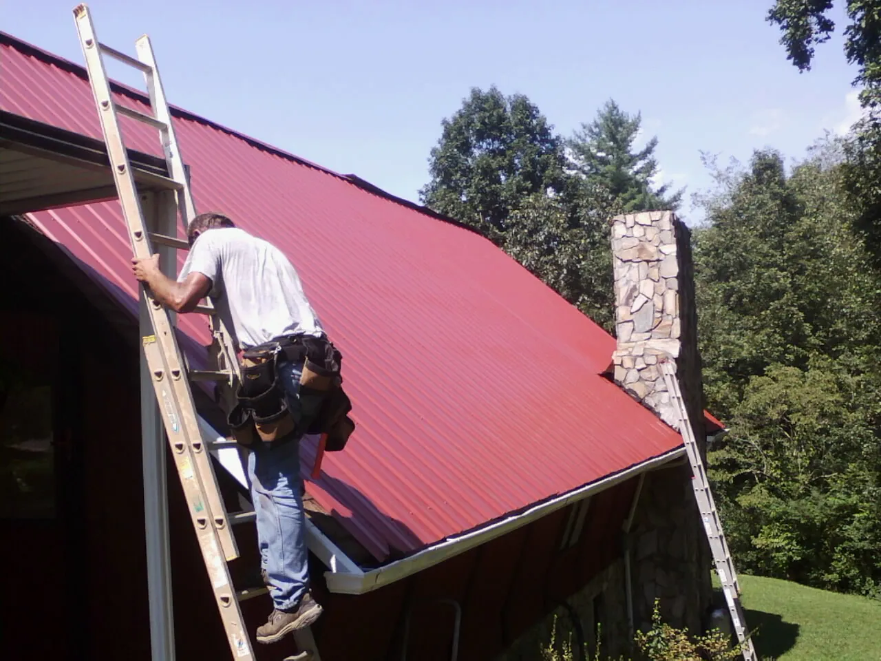 Roofer on a ladder beside a red metal roof and stone chimney — Soffit & Fascia Repair by Roofing Brothers in South Fallsburg